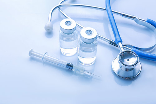 Medical supplies on a light blue background, including a stethoscope, syringe, and two sealed vaccine vials.
