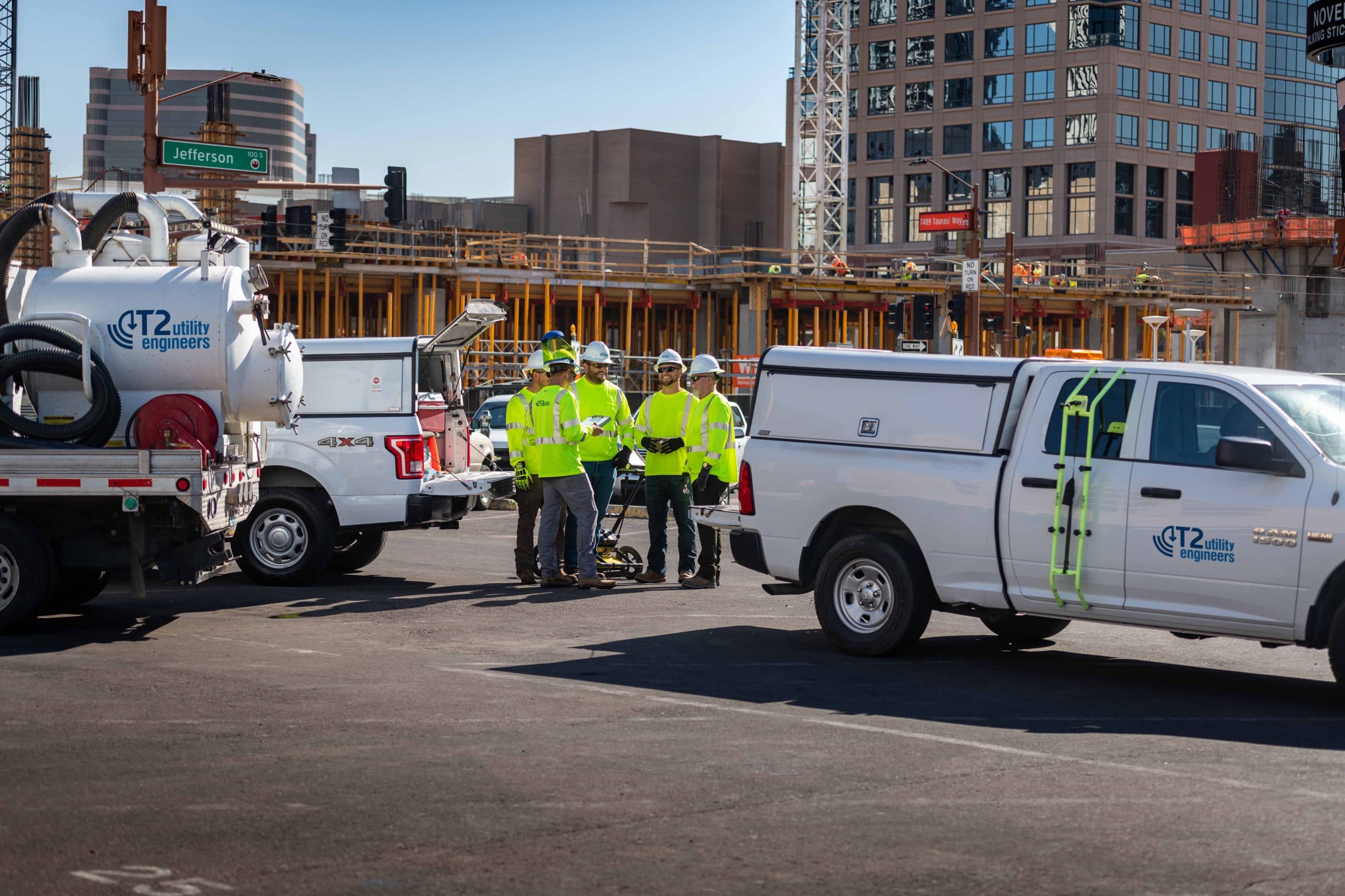 Kiwa T2 workers in safety vests and hard hats stand beside utility trucks at an urban building site.