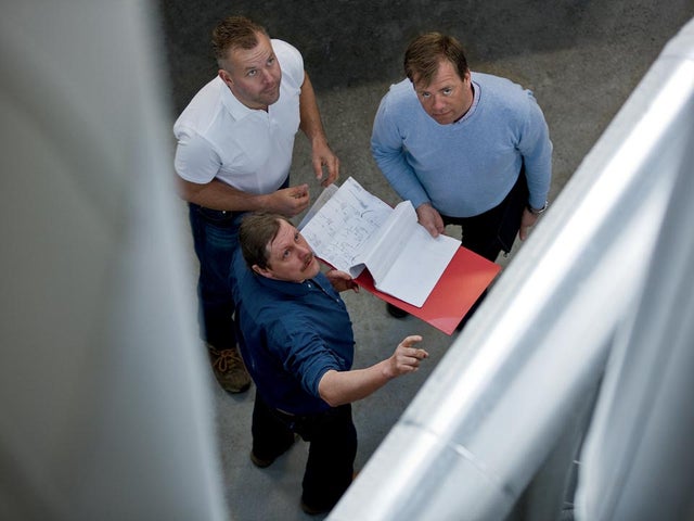 Three men standing together looking at open documents in an industrial or construction setting, viewed from above.