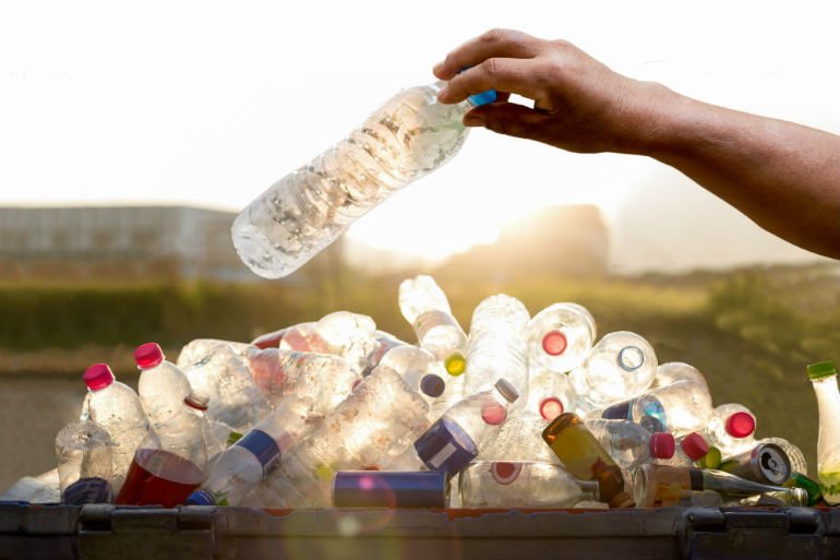 Hand holding a plastic bottle over a container filled with recyclable plastic bottles, set against a sunlit background