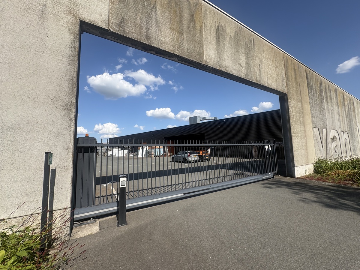 Moderne industriële ingang met een grote betonnen poort, metalen schuifhek en een afgesloten terrein onder een blauwe lucht met wolken.
