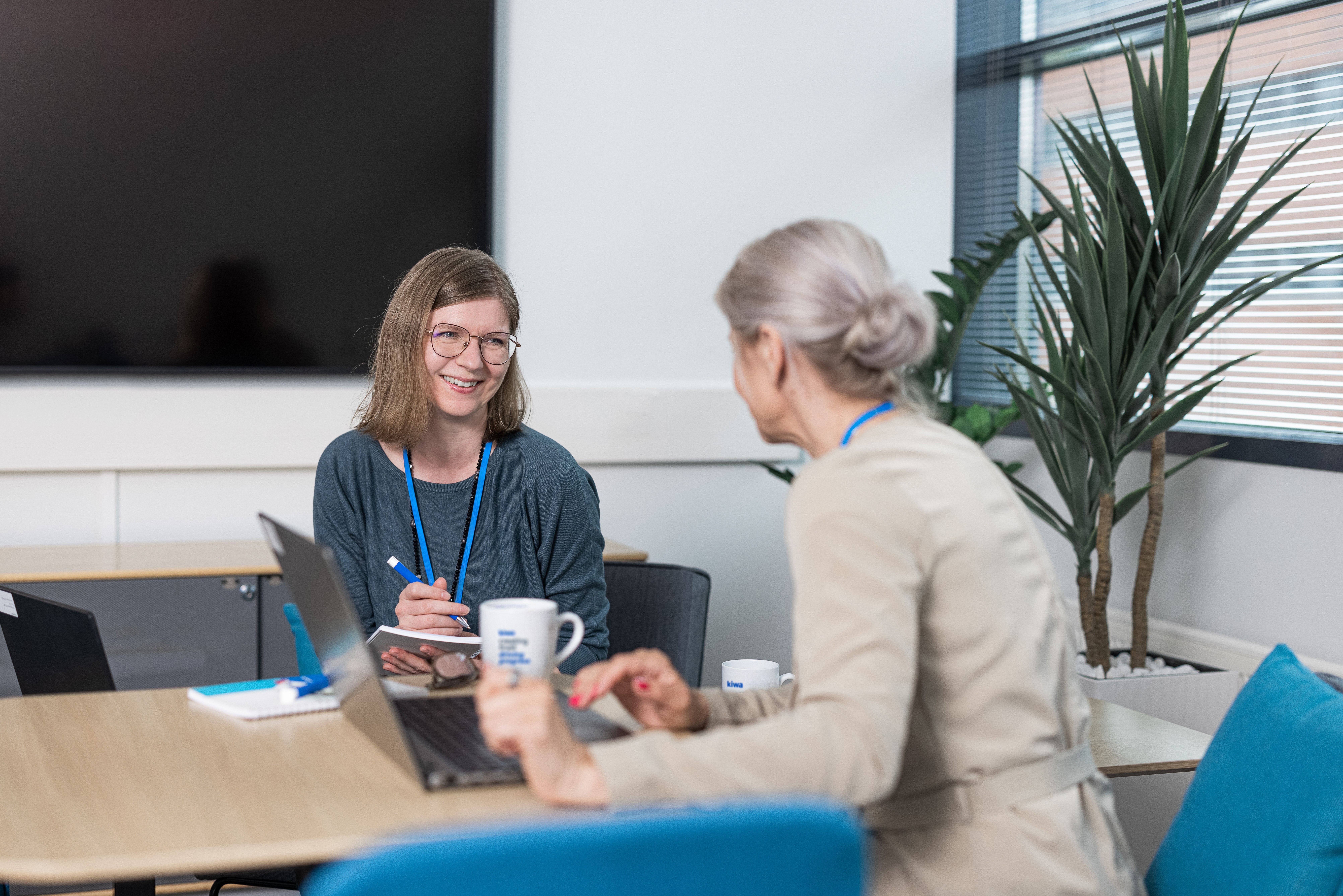 Two women in a business meeting at a conference table with laptops and coffee mugs, one taking notes while smiling.