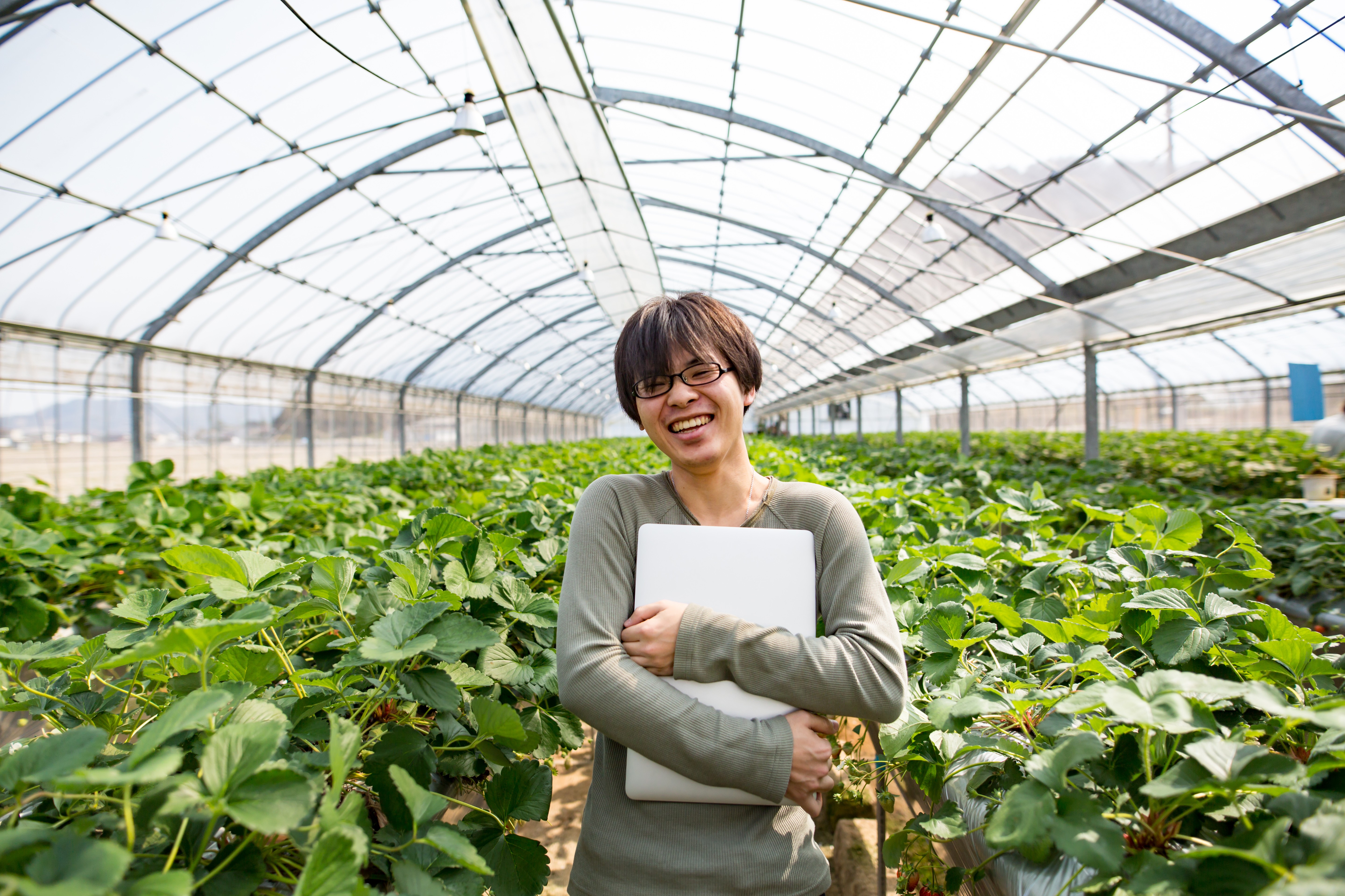 Portrait of a smiling woman holding a laptop in a greenhouse filled with green plants.