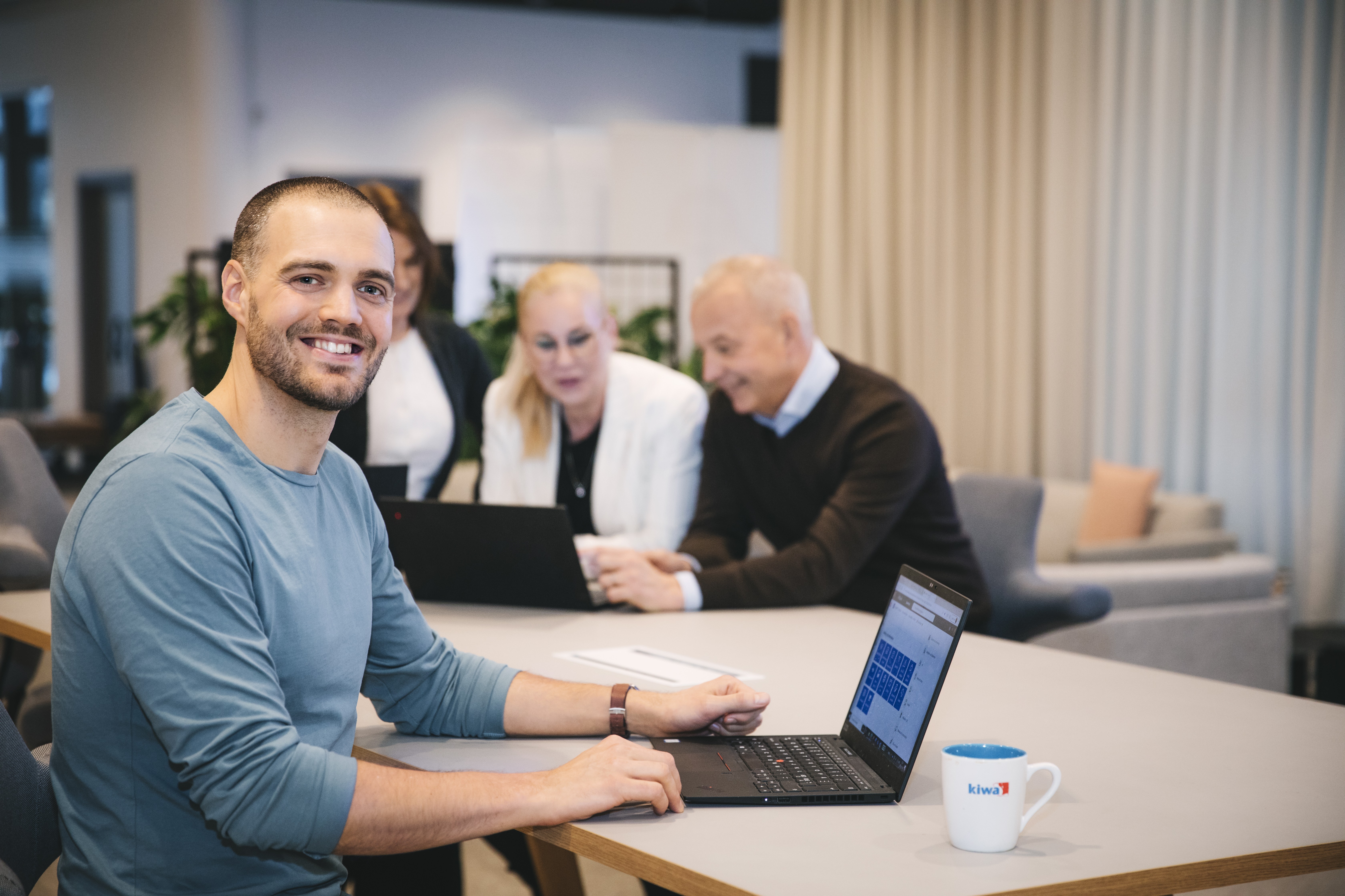 Man sitter vid laptop i kontorsmiljö med kollegor i bakgrunden. Affärsmöte med dator och kaffekopp på bordet.