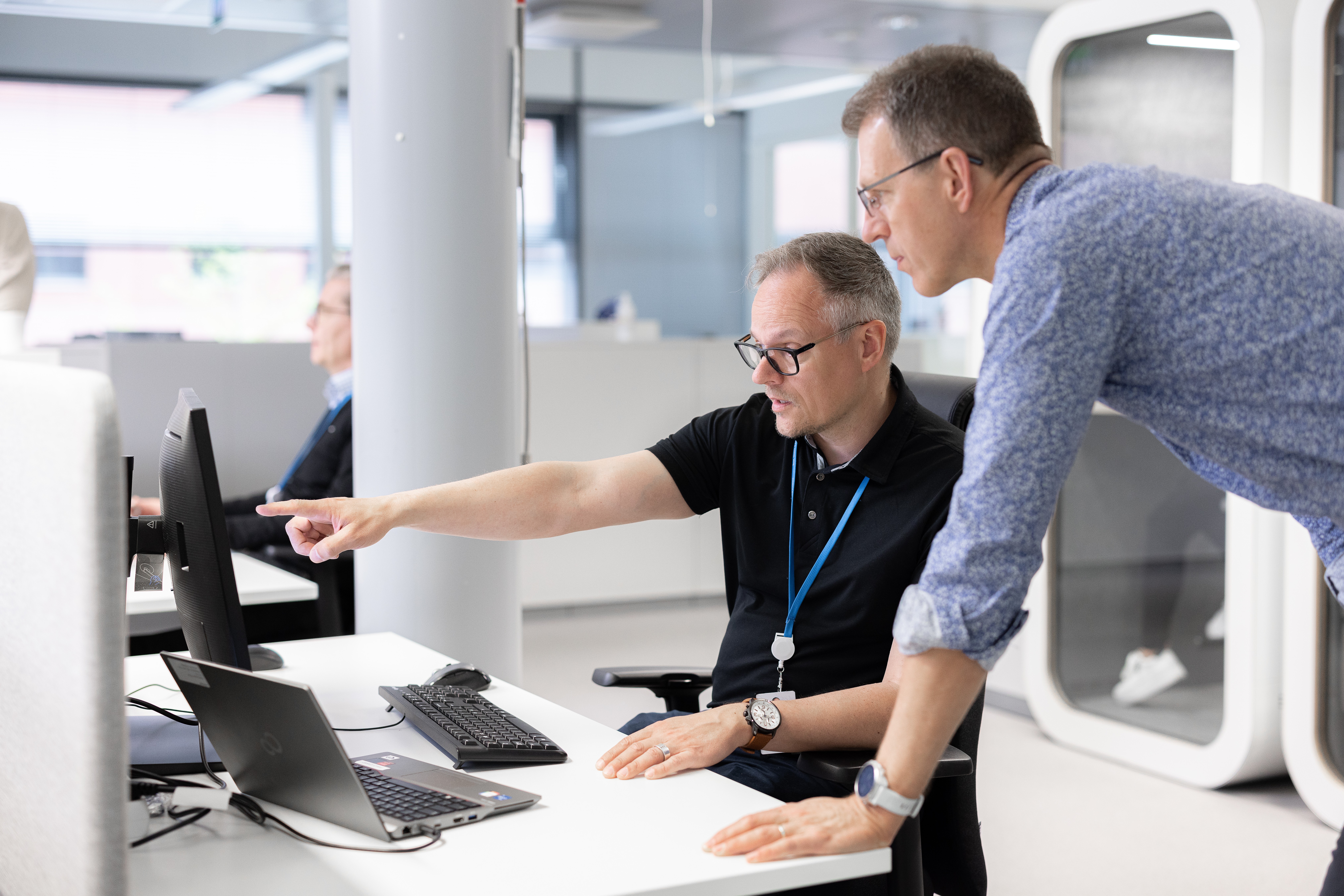 Two men in an office looking at a computer monitor, with one seated and pointing at the screen while the other leans over the desk.