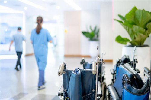 Hospital hallway with wheelchair in the foreground and two people in blue scrubs walking in the background.