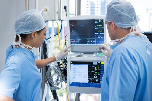 Two medical professionals in surgical scrubs and caps reviewing patient monitor displays in a hospital setting.