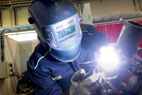 Welder wearing a helmet and safety gear, performing TIG welding on a metal pipe in an industrial workshop