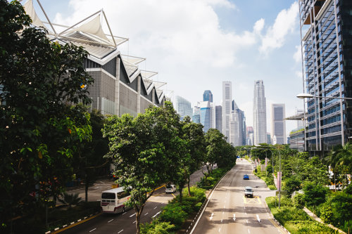 Ielas skats ar modernām ēkām, augstceltņu siluets un koki pilsētvidē saulainā dienā.