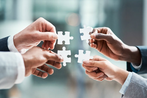 Mensen houden witte puzzelstukken vast tegen een achtergrond van een moderne glazen kantooromgeving.