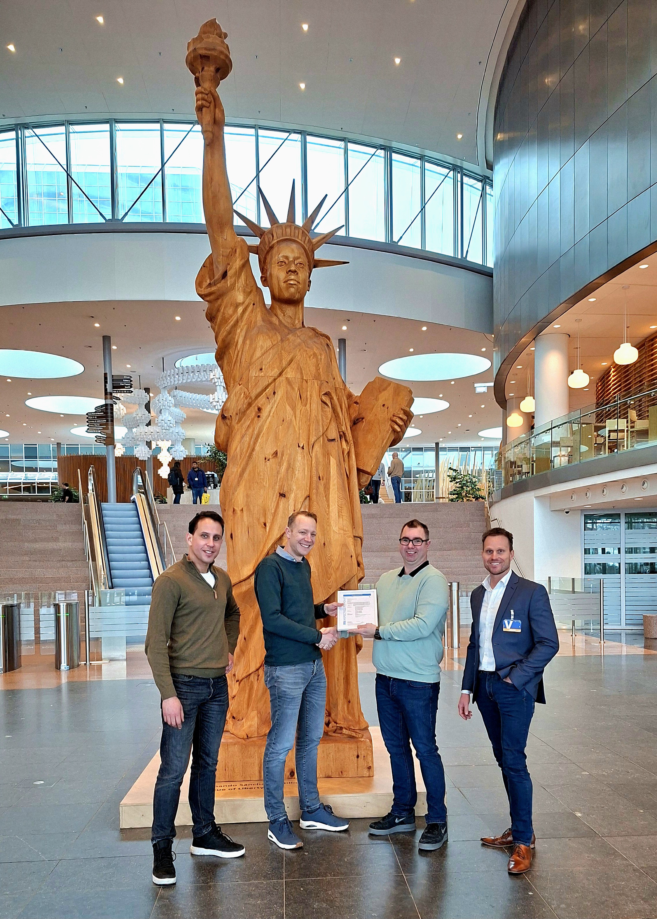 Vier mannen staan bij een houten replica van het Vrijheidsbeeld in een modern gebouw met glazen en metalen architectuur.