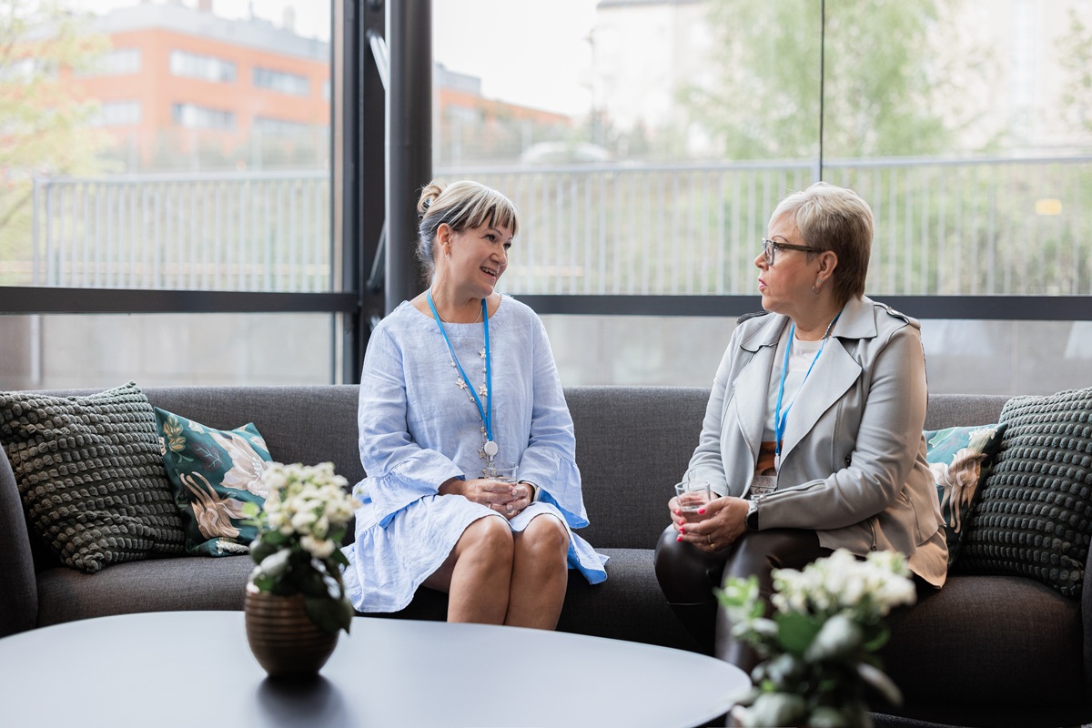 Two women talking on a sofa in a bright lounge, with lanyards and a coffee table in the foreground.