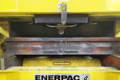 Gul hydraulisk press som böjer en räl i stål, så kallad böjprovning av räl som en metod inom förstörande provning, för att verifiera att rälen uppfyller ställda hållfasthetskrav.