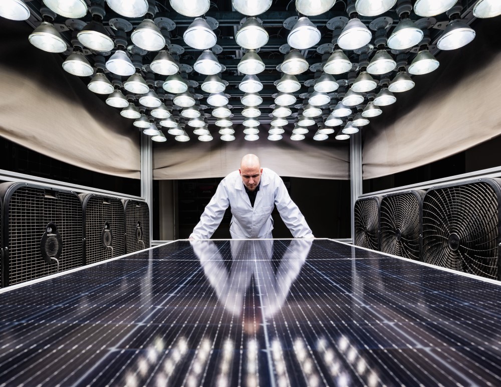 Kiwa PVEL laboratory employee in a white lab coat standing over a reflective solar panel under a grid of overhead lights, with large fans on both sides.