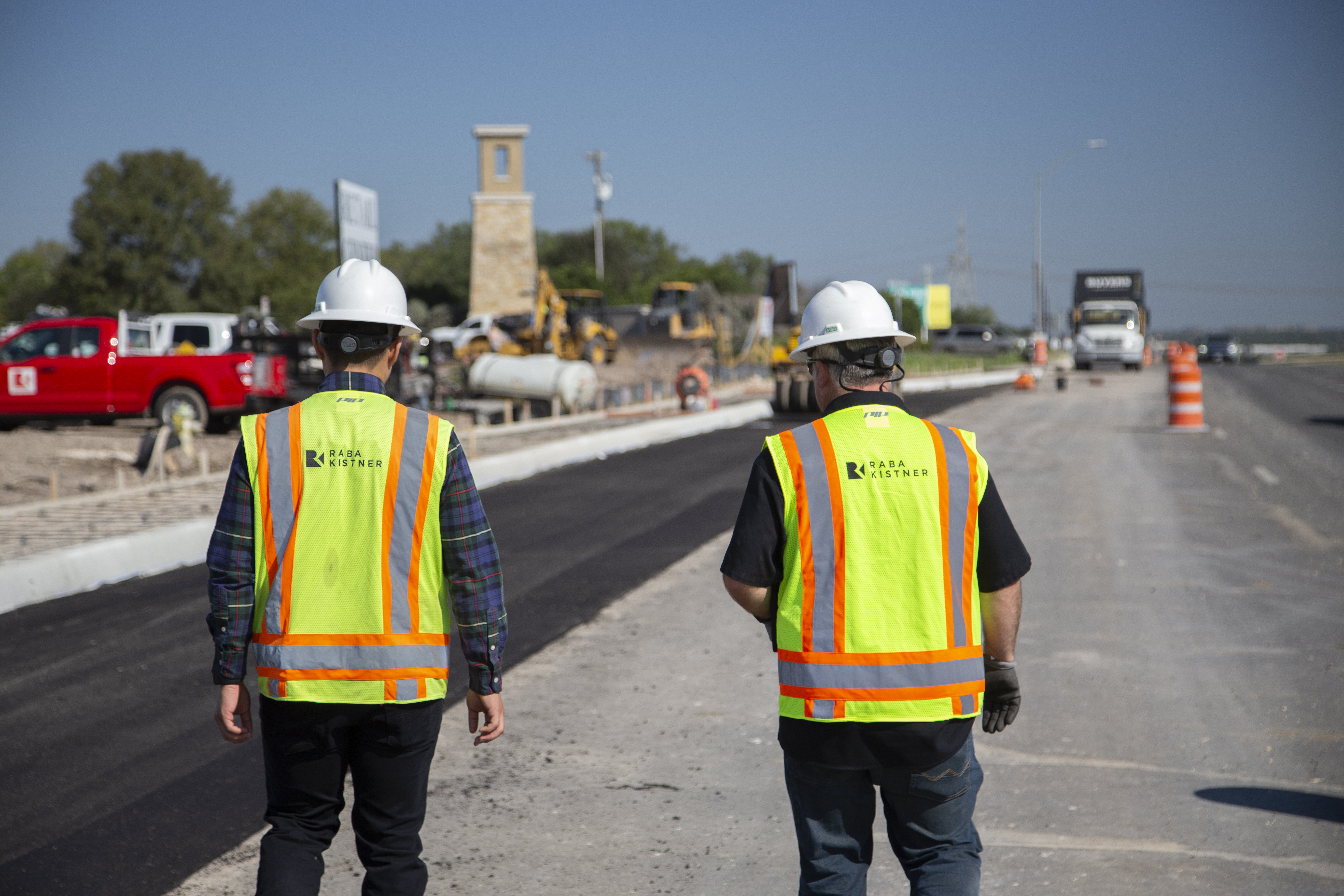 Two Kiwa employees wearing hard hats and high-visibility safety vests walk along a road construction site with asphalt paving equipment and traffic cones.
