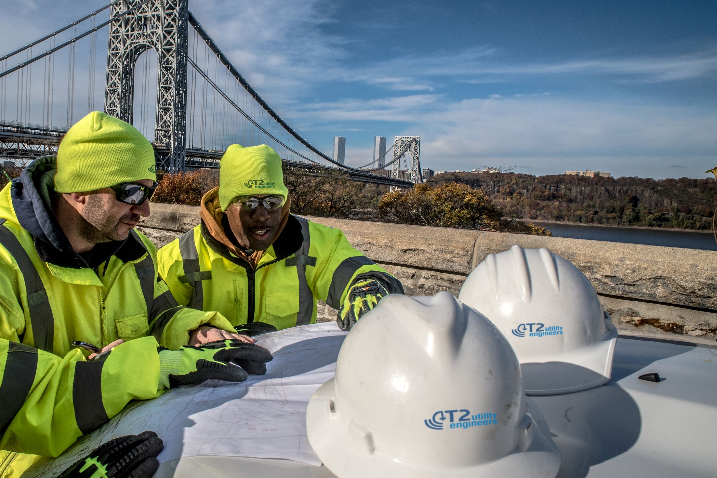 Kiwa utility workers in neon safety jackets and hats reviewing plans at a construction site with hard hats on a table and a suspension bridge in the background