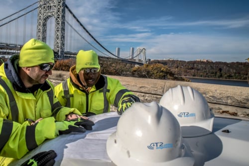 Kiwa utility workers in neon safety jackets and hats reviewing plans at a construction site with hard hats on a table and a suspension bridge in the background