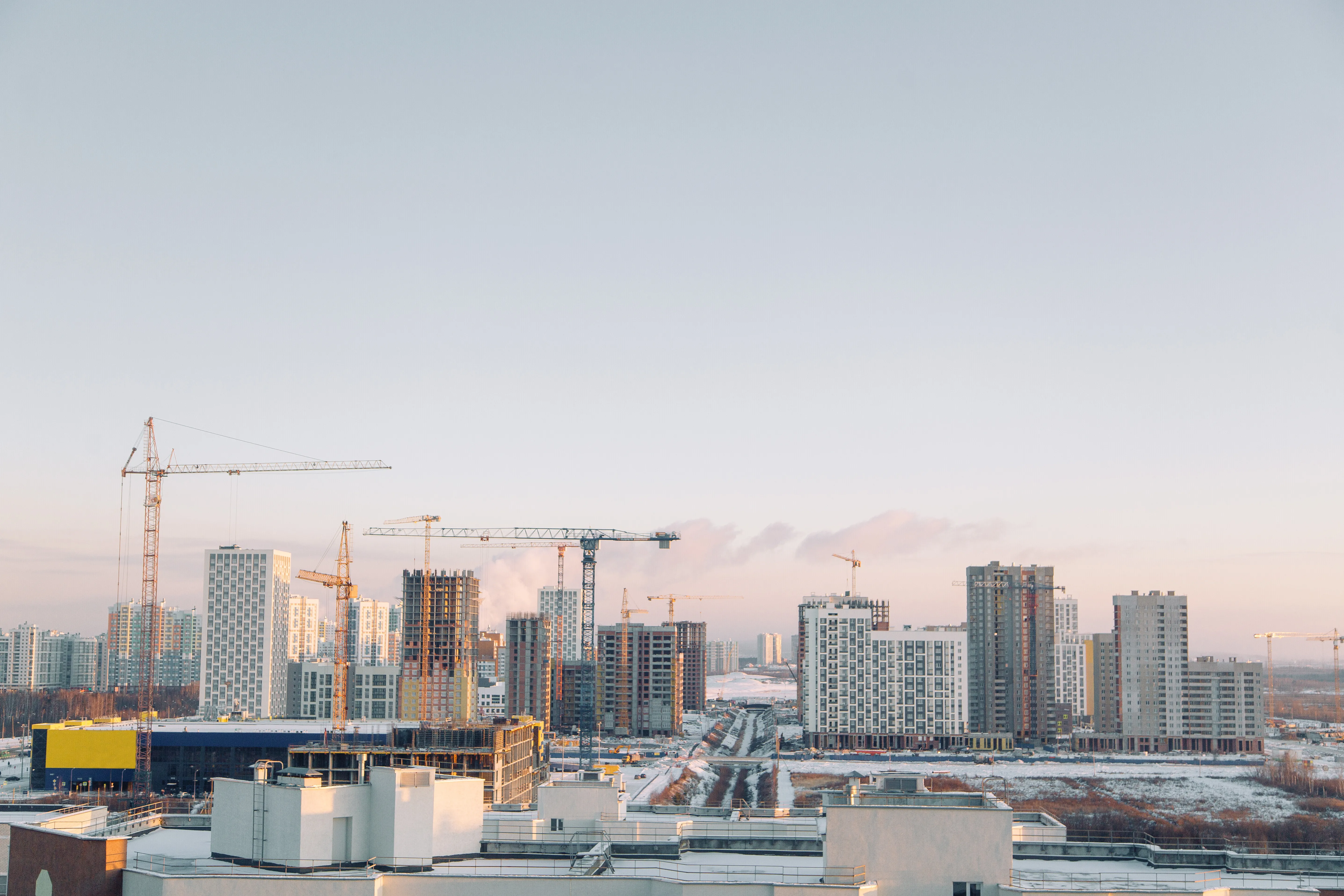 Urban skyline with modern apartment buildings, construction cranes, and a snowy landscape under a clear sky.