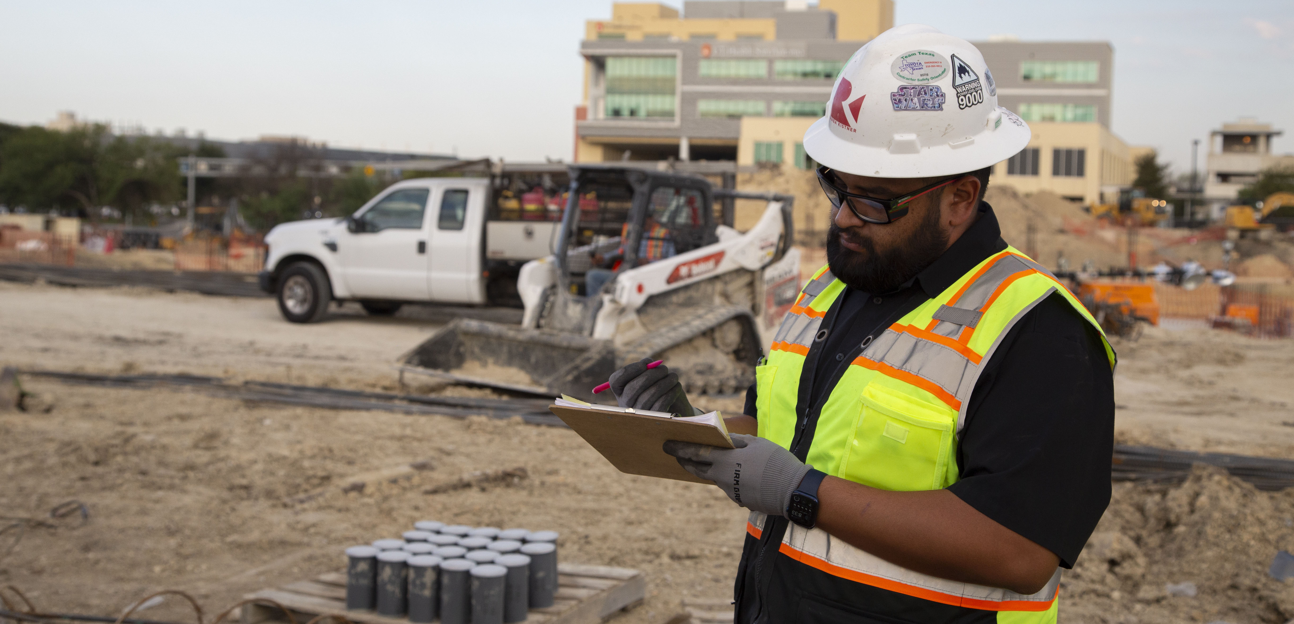 Raba Kistner employee in a hard hat and safety vest taking notes at a building site with a pickup truck and skid steer in the background.