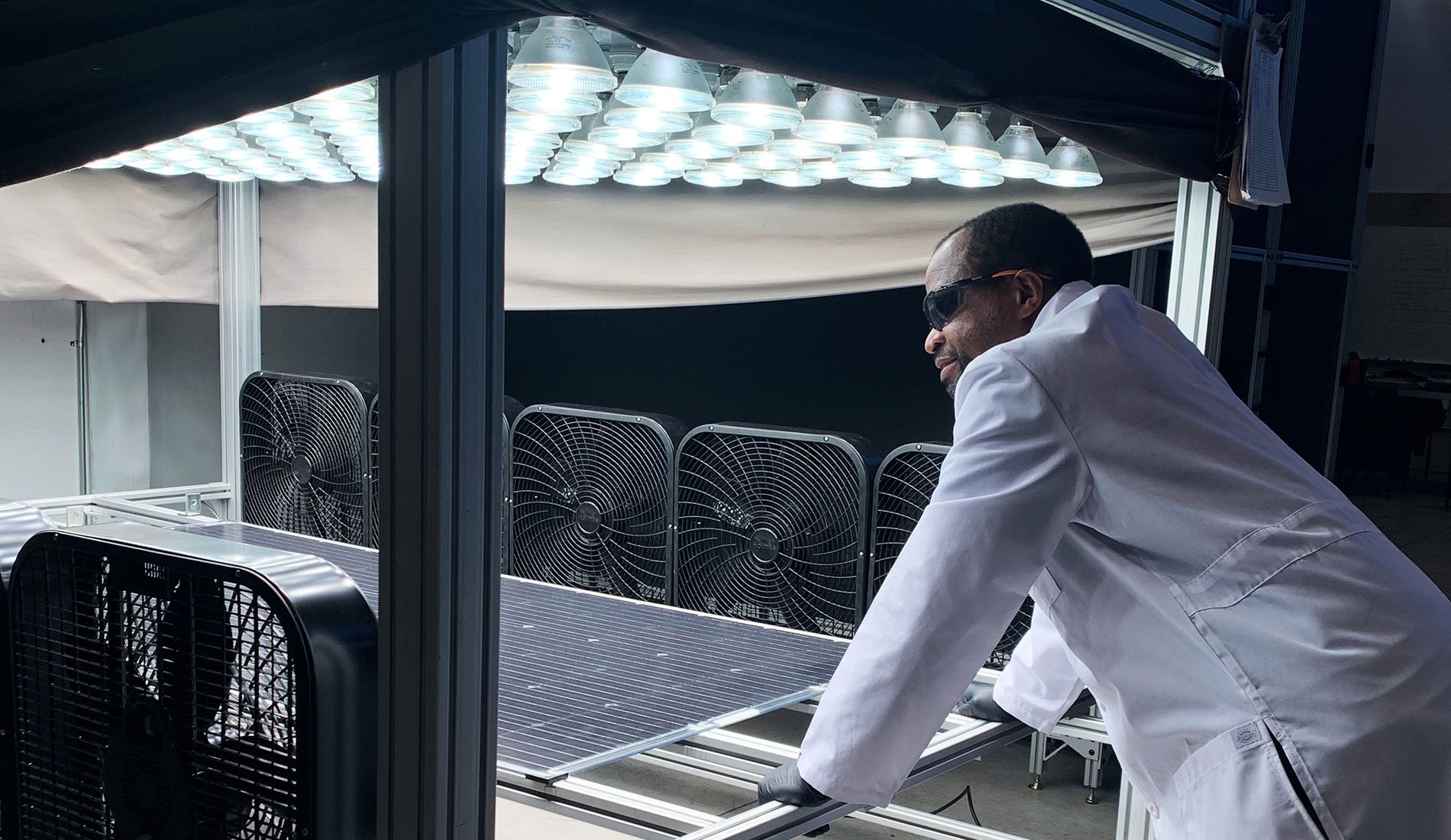 A Kiwa lab employee in a white lab coat examining solar panels under an array of artificial lights with multiple fans in the background.